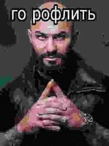 a bald man with a beard is sitting with his hands folded in front of him .