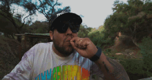 a man wearing sunglasses and a tie dye shirt is smoking a cigarette outdoors