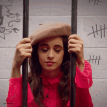 a woman wearing a beret is behind bars with a brick wall behind her that has a drawing of a skull