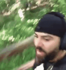 a man with a beard wearing headphones and a beanie is walking through a forest .