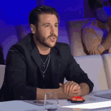 a man with a beard is sitting at a table with a microphone and a glass of water .