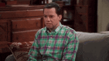 a man in a green plaid shirt sits on a couch