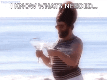 a man with a beard is standing on a beach holding a glass of wine and saying `` i know whats needed ... ''