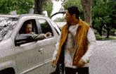 a man in a brown vest is talking to another man in a silver suv