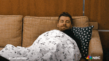 a man is wrapped in a blanket on a couch with a nbc logo in the corner