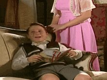 a young boy is sitting on a couch reading a book while a woman in a pink dress holds his hair .
