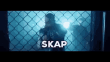 a man is standing behind a chain link fence in a dark room with the word skap written above him .