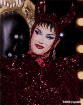 a drag queen is sitting on a throne wearing a devil costume and sequins .