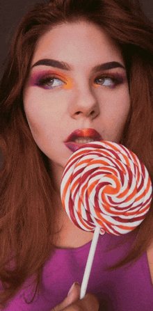 a woman in a purple top is holding a colorful lollipop in her mouth