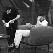 a man is sitting in a chair while another man pours something into a cup