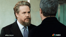 a man with a beard is standing next to another man with a nbc logo in the corner