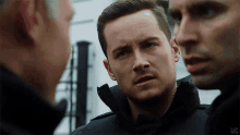 a man with a beard is talking to two other men while wearing a black jacket .