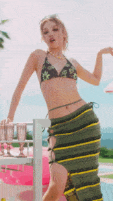 a woman in a bikini and a striped skirt is standing next to a table with glasses on it .