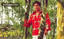 a man in a red plaid shirt is standing next to a tree in the woods .