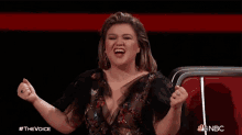 a woman in a black dress is screaming with her fist in the air and the words #thevoice nbc behind her