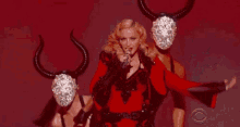 a woman in a red dress is singing into a microphone while a man in a mask with horns holds her .