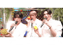 three men singing into microphones while holding fruit including a mango