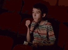 a young boy in a striped shirt is sitting in a movie theater eating popcorn
