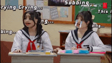 two girls are sitting at desks in a classroom and one of them is crying and the other is bing