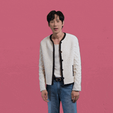 a man wearing a white cardigan and blue jeans stands in front of a pink background