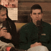 a man in a green jacket is talking into a microphone in front of a calendar that says march 2025