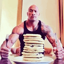 a muscular man is sitting at a table holding a fork and knife next to a plate of pancakes .