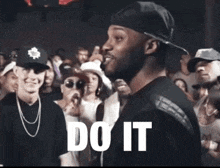 a man in a hat is standing in front of a crowd with the word do it written on his chest .