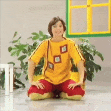 a young boy in a yellow shirt and red pants is sitting on the floor in front of a window .