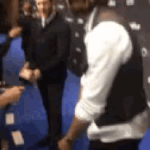 a man in a vest is standing on a blue carpet talking to another man .