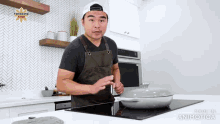 a man in an apron is standing in a kitchen with a pan on a stove top .