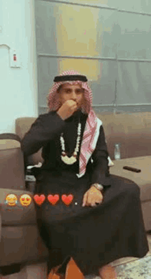 a man in a keffiyeh is sitting on a couch with his hand to his mouth .