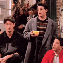 a man in a coat is holding a cup of coffee while sitting in a room with other people .