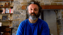 a man with a beard wearing a blue shirt is sitting in front of a fireplace