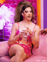 a drag queen is sitting on a couch holding a drink
