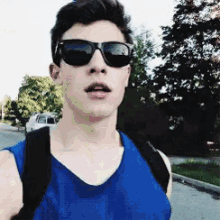 a young man wearing sunglasses and a blue tank top looks at the camera