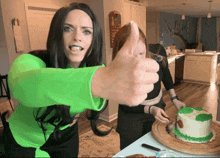 a woman in a green shirt gives a thumbs up in front of a cake
