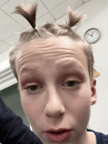a boy with pigtails on his head looks at the camera in front of a clock that shows the time as 4:20