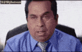a man in a blue shirt and tie is sitting in a chair and making a funny face .