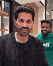 a man with a beard is smiling while standing next to another man in a green sweater .