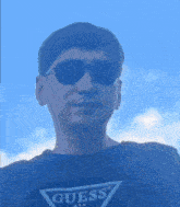 a man wearing sunglasses and a guess shirt looks at the camera