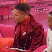 a man in a red jacket and turtleneck is sitting on a pink couch holding a piece of paper .