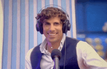 a man wearing headphones and a vest smiles in front of a microphone