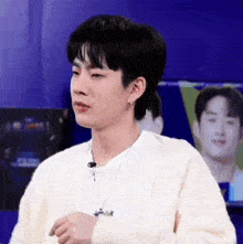 a young man wearing a white sweater and a necklace is standing in front of a television .