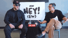 two men are sitting in front of a sign that says hey ( ew )