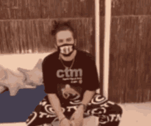 a man wearing a mask and a ctm shirt is sitting on a bed
