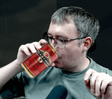 a man wearing glasses is drinking from a red can that says ' britannia ' on it .