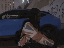 a man with dreadlocks is standing in front of a blue car with graffiti on it .