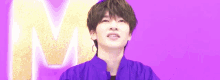 a man in a purple jacket is standing in front of a purple background with a large m in the background .