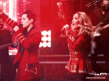 a man in a red jacket sings into a microphone next to a woman in a red dress with her arms in the air