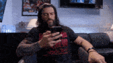 a man with long hair and a beard is sitting on a couch looking at his phone
